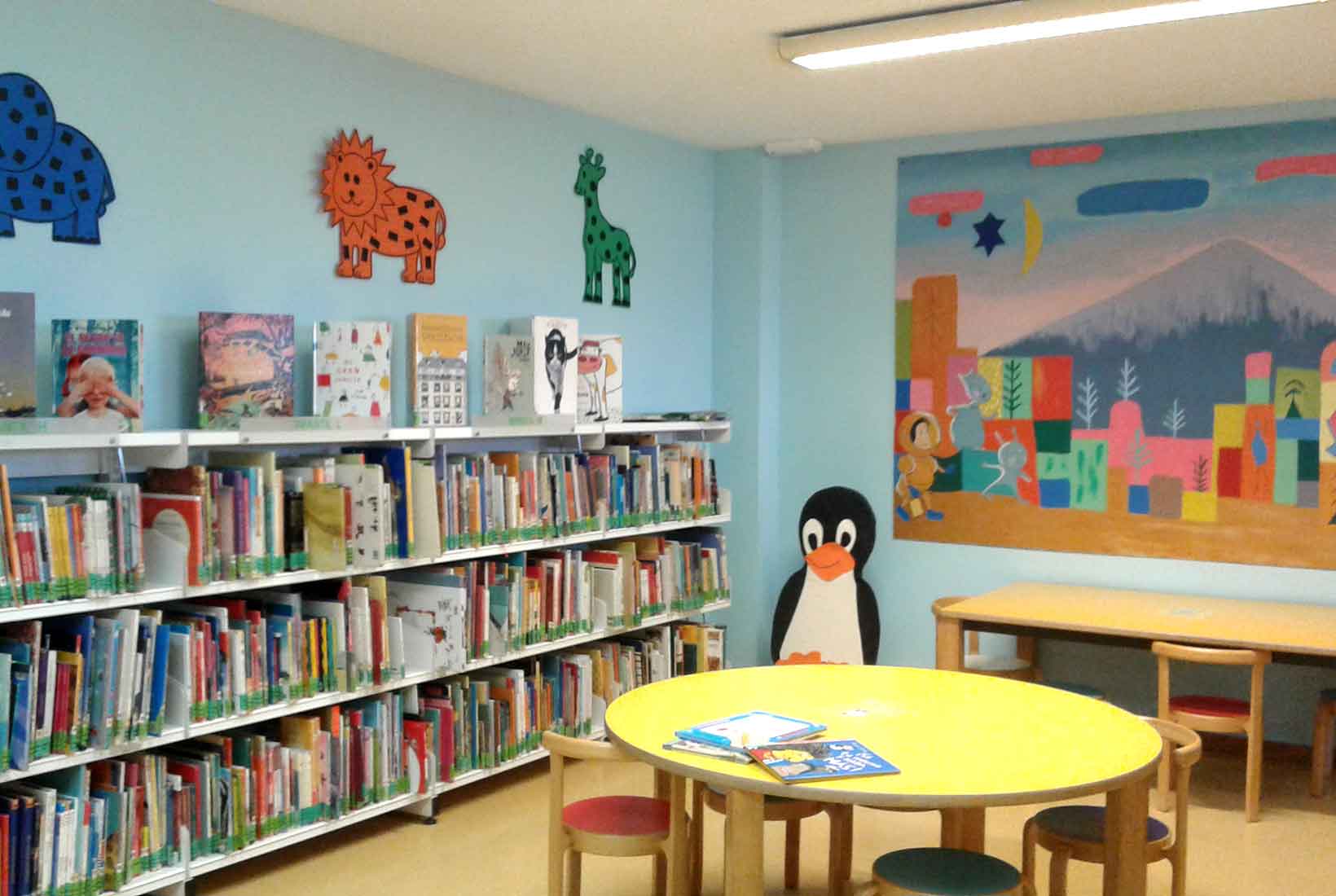 Sala de la biblioteca pública de León con varias mesas y sillas bajas,una estantería llena de libros infantiles y paredes azules decoradas con dibujos. 