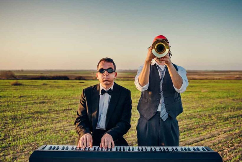 Un hombre con gafas de bucear toca un teclado y a su lado otro toca la trompeta con un fondo campestre