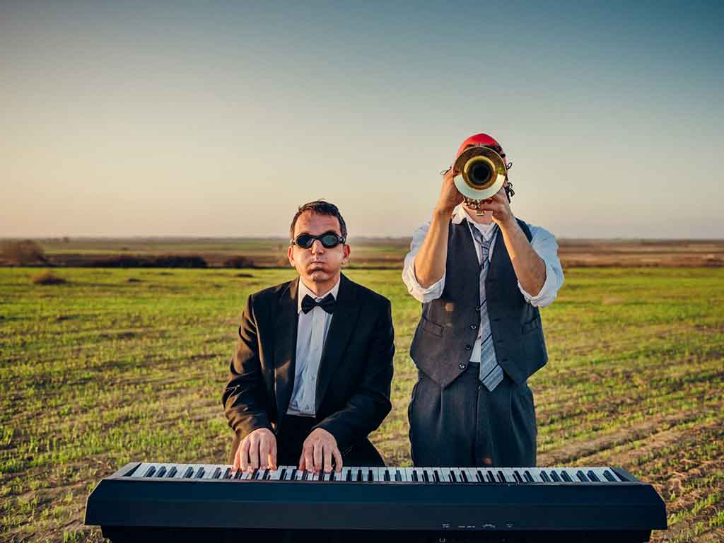 Un hombre con gafas de bucear toca un teclado y a su lado otro toca la trompeta con un fondo campestre