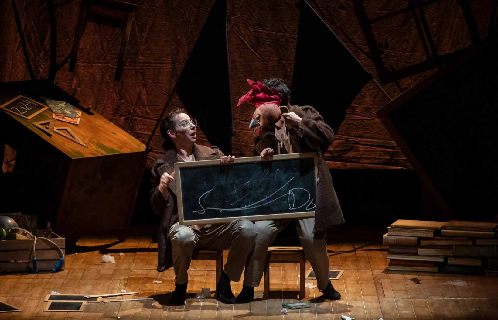 Una mujer se sorprende de una especie de hombre pájaro mientras ambos sostienen en una pizarra en un escenario que simula una escuela. Es una escena de la obra de teatro familiar La Maestra.