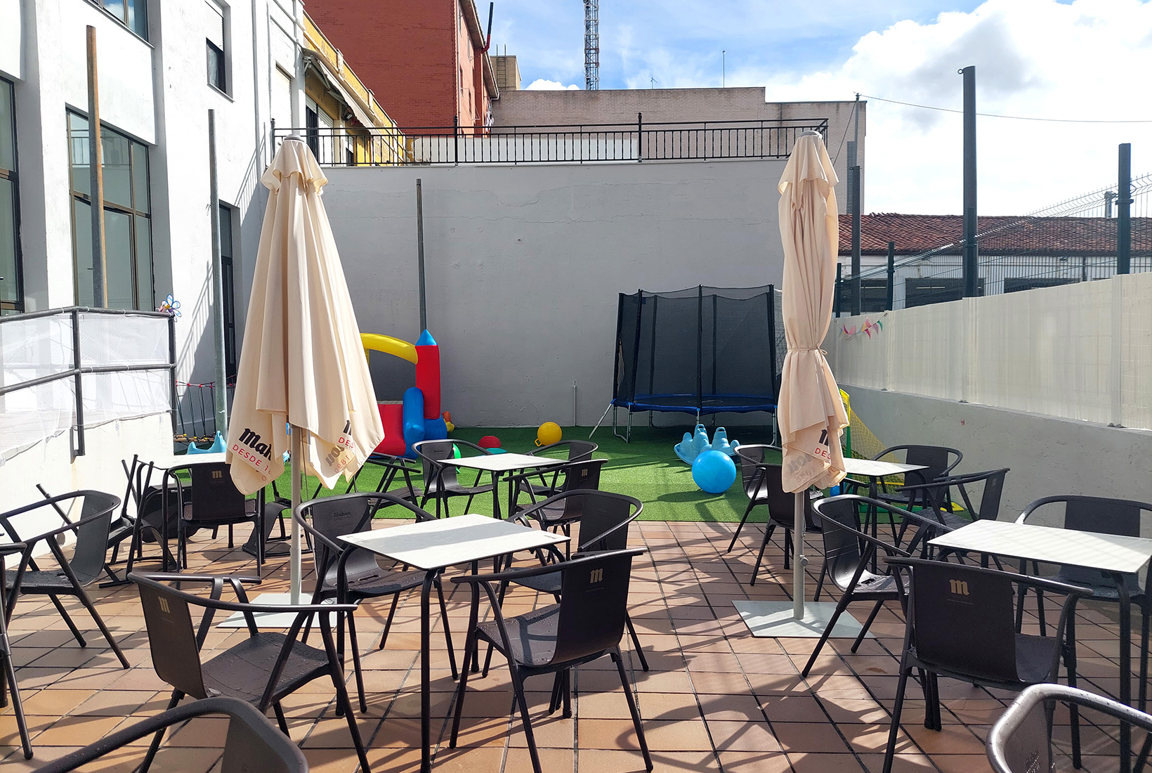 Terraza con varias mesas y sillas y sombrillas en Limalimón, una cafetería para familias en León. Al fondo se ve un pequeño hinchable, una cama elástica y otros juegos por el suelo.